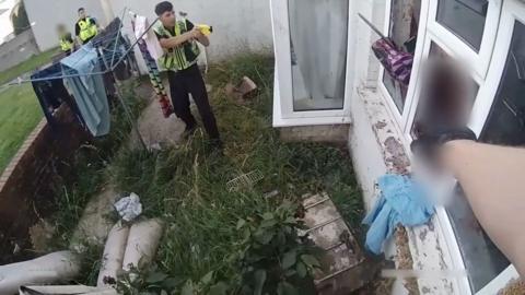 A man reaches for something near a window. Two officers can be seen on the left. One is aiming his taser at the man.
