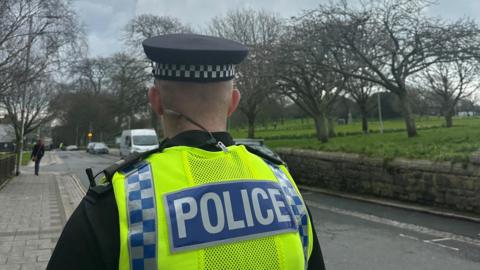 A man, facing away from the camera. He is in police uniform and a police hat. The road is to the right of him and he is stood on the pavement. To the right of the road there is a wall and grass and trees. 