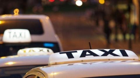 A stock image of a row of taxis, heading into the distance away from the camera. Only the tops of the taxis are visible, their signs are illuminated.