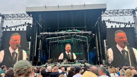 A picture taken from the crowd of three screens showing Bruce Springsteen playing on stage. Springsteen has short grey hair and is singing into a mic, he is wearing a white shirt, black tie and waistcoat.