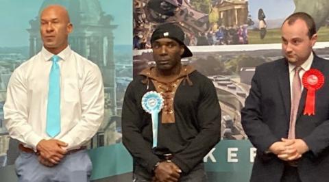 Three men at a local election for Wakefield in May 2026 - a tall, bald man is on the left wearing a white shift, turquoise tie and blue trousers; the man in the centre has a black baseball cap on backwards and is wearing a black and brown hoody with black jeans; the man on the right has short black hair, a black suit and a pink tie. All three men have their hands clasped in front of them.