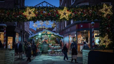 A festive street scene at what appears to be a Christmas market, captured during the evening or early night. A large decorative arch spans the street in the foreground, lush with green garlands, baubles in red and gold, and warm white lights. Several glowing star-shaped light decorations hang from the arch, creating a welcoming entrance and framing the view beyond. Underneath the arch sits a wooden market stall, topped with greenery and lights. The stall is filled with seasonal items—likely Christmas ornaments, wreaths, plants, and small festive decorations—arranged neatly on shelves and tables. The street is narrow and lined with brick buildings, their shopfronts softly illuminated. People in winter coats and scarves walk through the scene, some browsing the stall, others passing by with shopping bags. 