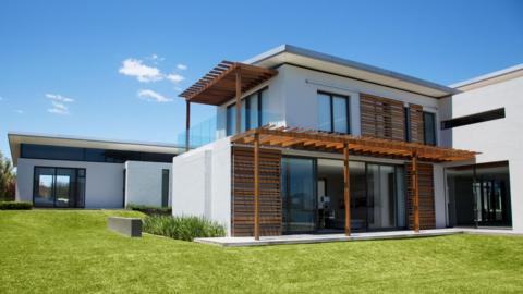 A large modern modern house with whitewashed walls and wooden trimmings. Large glass doors lead out onto a grass lawn. Above the house is a blue sky with a couple of clouds. 