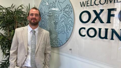 Mr Bearder is wearing a white suit and silver tie. He is standing next to the Oxfordshire County Council logo.