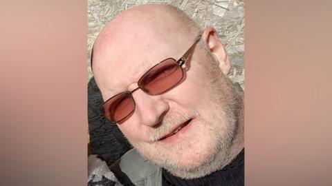 A bald man with a white beard and brown-tinted sunglasses inclines his head as he looks towards a camera. He is outside on a sunny day.