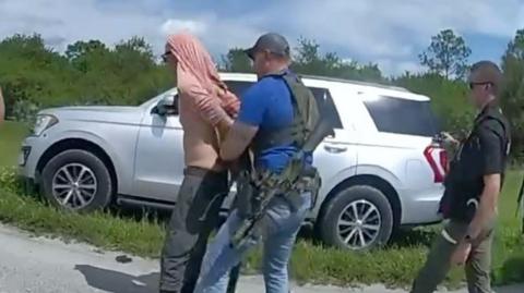 A man with a pink shirt over his head is handcuffed.