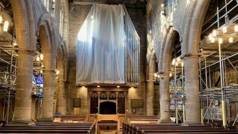 Interior of church with massive white drape covering a walled section and metal scaffolds around the sides. Pews and vaulted wooden arches in front of stained glass windows.