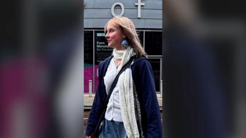 Young woman with blonde hair, looking to her right with a smile in a street.