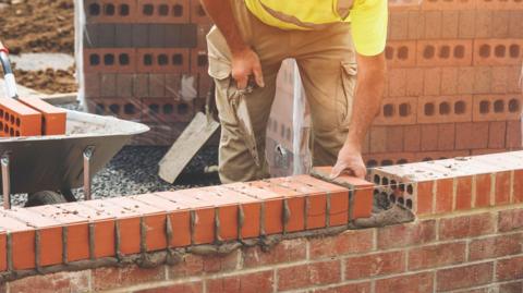 A builder wearing beige cargo trousers and a yellow high-visibility top is placing a brick into a wall which is under construction. He is holding a trowel and is leaning forwards over the wall.