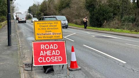 Image shows a road with traffic. There is a sign and a traffic cone saying Road Ahead Closed, and a yellow information sign saying that Horsebridge Hill is closed from Nicholson Street and that this road (Horsebridge Hill) is a no through road.
