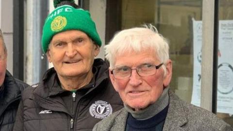 Two men pictured outside the trophy shop.  Gerry Doc has a green hat and black and green jacket, John 'Jobby' Crossan in a grey suit and jumper