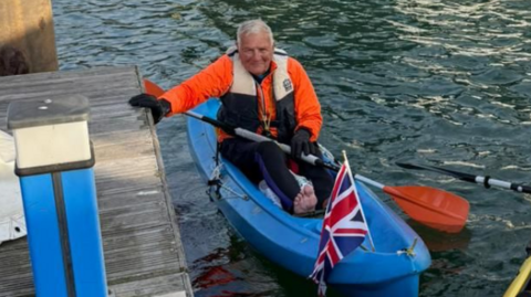 Barry Samples sits in a blue kayak on the water. He is wearing a life vest over a bright orange jacket and dark-coloured wetsuit. He is holding a paddle in one hand and the other is resting on a wooden platform. There is a red, white and blue Union Flag stuck to the front of the kayak.
