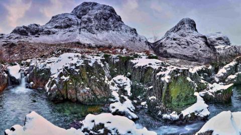 Water flowing between rocky ground covered in snow with the Three Sisters of Glencoe in the distance