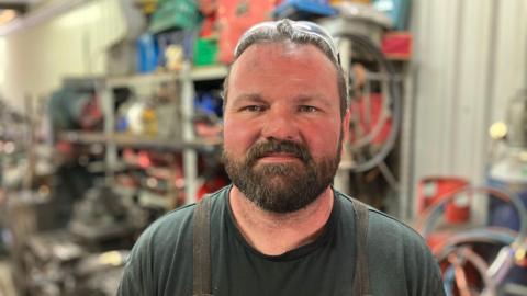 Ben Cooper, a man wearing a dark green t-shirt and an apron, is stood in a blacksmith's forge.