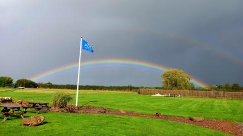 One very vivid rainbow stands out against a grey sky over a very green field. There is also a fainter rainbow in the sky above it. In the foreground there are two wooden picnic benches on the left and a flag pole with a blue flag attached.