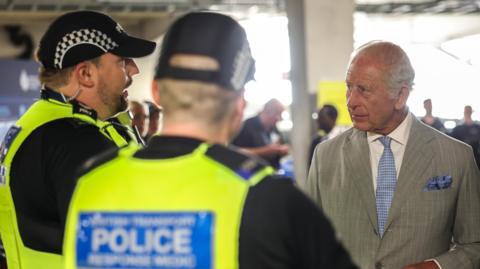 King Charles III wearing a light grey suit speaks with two police officers wearing high-visibility uniforms inside a large indoor venue. The officers stand in the foreground. Bright lighting and a shallow depth of field draw attention to the interaction. The background contains several blurred figures and structural elements of the building.