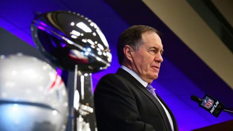 Bill Belichick, pictured in 2019, talking into an NFL-branded microphone at a press event. He has short dark hair and is wearing a dark suit with a pale short and purple tie. To gis right are the Super Bowl trophy and a New England Patriots helmet.