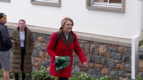 Deputy Sasha Kazantseva-Miller - A woman with blonde hair walking up a hill, wearing a red coat and a back pack. In her right hand she is carrying a green carrier bag. 