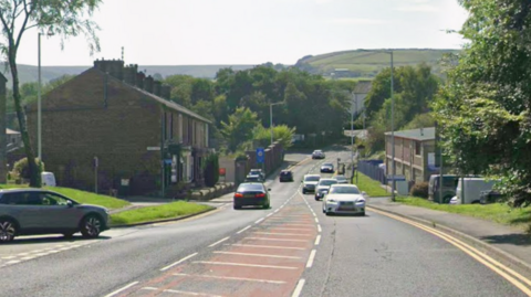 Google streetview of Grane Road, with cars on the carriageway. Terraced houses line the left of the road and trees on the right. Green hills can be seen in the backdrop.