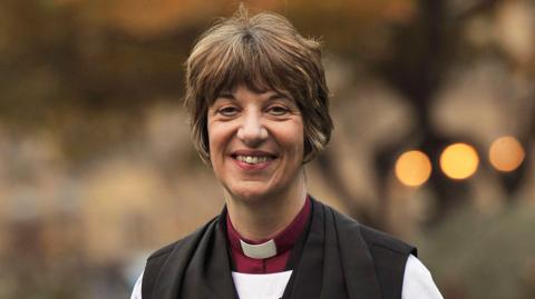 The Bishop of Gloucester, the Right Reverend Rachel Treweek, looks at the camera, smiling, with short, dark hair and with the typical clergy dog-collar and robe round her shoulders.