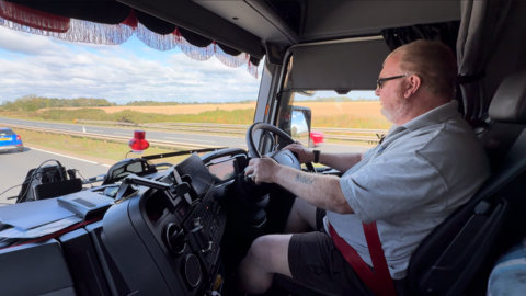 Man behind the wheel of a truck. he is sat up high and cars are on the road next to and in front of his cab.