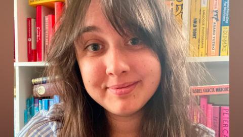 A young woman with long brown hair, wearing a blue and white striped shirt, smiles at the camera in front of a shelf lined with books. 