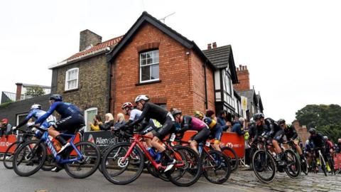 A group of cyclists arrive at the top of Michaelgate in Lincoln. The riders are all wearing lycra and cycling helmets as they travel over a cobbled road. A period building is visible in the background.