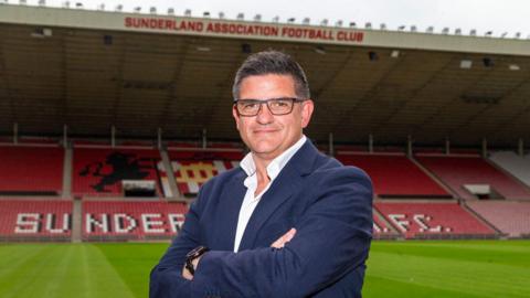 James Gray, wearing dark suit and white shirt with open collar, stands with arms folded in front of a large stand at Sunderland's Stadium of Light home