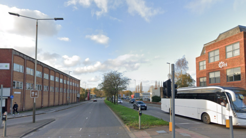 A dual carriageway road with trees along a grass central reservation.  A white bus travels along the right-hand side of the image. The road is lined by office and industrial properties.