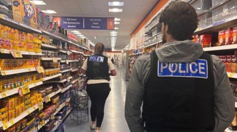 A man police officer- wearing a black police vest with police written in blue and a grey hoodie and a woman officer also wearing the black police vest and a white short-sleeve T-shirt and black leggings- walking inside supermarket aisle.