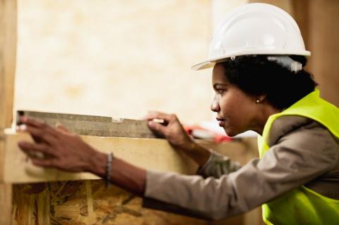 A woman in a hard hat wears a green high-vis jacket over a grey top while looking at a slide rule, a metal bar, on top of a wooden lintel in a window. 