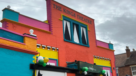 The facade of the theatre which is very brightly painted in a variety of bold colours, including red, yellow, blue and pink. There are green, gold and white balloons tied to the building