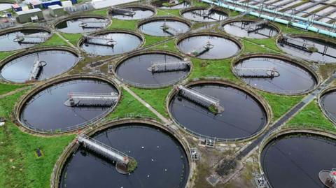 A general shot of a water treatment plant. There are several circular vats of water, each with an arm stretching from the middle to the edge of the circle. In between the circles are patches of grass.  