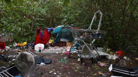 An encampment on a muddy clearing in a wooded area, including a tent, litter strewn around and two shopping trolleys. There are trees surrounding the area.