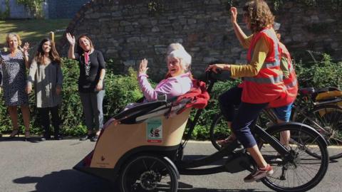 Two elderly ladies sit in the front carriage of a tricycle which is being ridden by a younger woman. People standing nearby wave at them. 