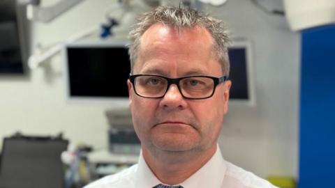A man in a hospital setting. He is wearing black rectangular glasses and is wearing a white shirt. He has a short grey hair and is staring into the camera. 