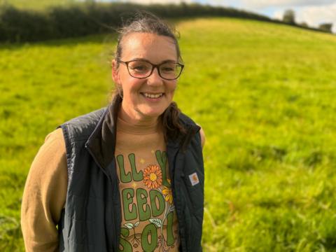 Bronagh O'Kane, standing in a field, with shoulder length dark hair, wearing glasses. She is smiling at the camera wearing a black gilet over a yellow jumper