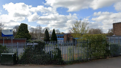 A school behind grey metal fencing with a name sign to the left and a blue-framed building in the distance