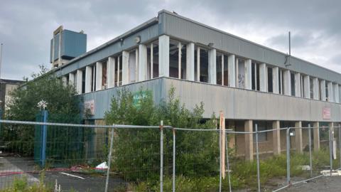 Abandoned school building behind fencing, with broken windows and graffiti.