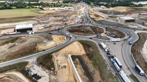 Aerial view of roadworks around a large roundabout with traffic queuing both southbound and northbound. Either side of the roundabout workers are building the beginnings of a flyover.