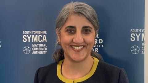 A woman with grey hair is smiling and wearing a navy blue suit with yellow trim. She is stood in front of a blue board which says South Yorkshire SYMCA in white writing