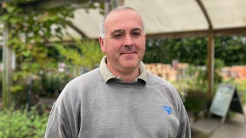 A man with short grey hair, wearing a grey jumper. There are plants behind him