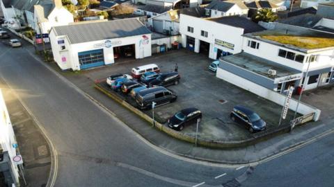 Tower Road Garage pictured. The site is comprised of three buildings. The first had large shutters and just one storey. The second has two storeys with two large doors on the ground floor and two small windows on the second floor. The third building, to the second building's left, is again two storeys. It appears to have a shop frontage facing the road. Each building has been painted white. There is a large forecourt with several vehicles parked on it.