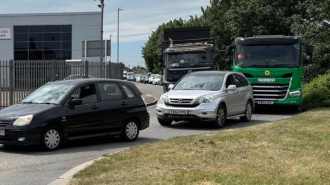 Cars and trucks of various colours queuing in a narrow street on an industrial estate. The queue stretches back as far as we can see. In the foreground is a patch of green grass. In the background are green trees.