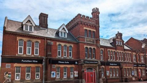 Red brick Victorian building with rectangular windows and a red front door - its roof in the centre has a castle-style turret.