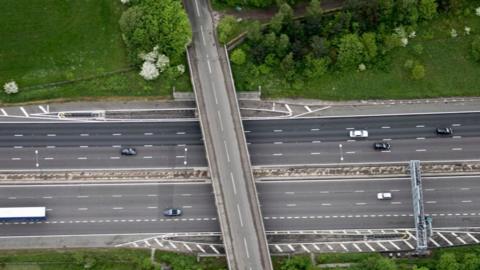 A birds-eye view of a motorway. There is a bridge running across it. Trees/ a forest at the top of the image. 