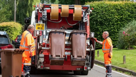 Two men in orange hi-viz overalls stand either side of the back of a waste collection vehicle. The truck is in the process of emptying two brown bins. Behind the van there is a patch of grass and several large green bushes, as well as a couple of parked cars on the road.