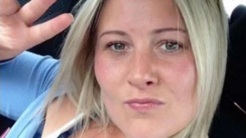 Joanne Penney seen in selfie taken sitting in a car. She is making the peace sign hand gesture and has shoulder-length straight blonde hair and wears a light blue cardigan.