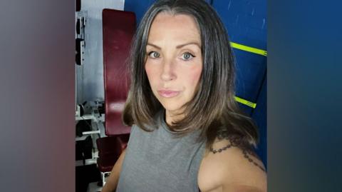 Jacqui Oakes wearing a grey vest at the gym. She has long brown hair past her shoulders and behind her is a piece of gym equipment.