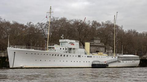 The portside view of the HMS Wellington, which is docked on a river on Victoria Embankment on an overcast day. It is a large, white vessel. There are trees behind it on the bank.